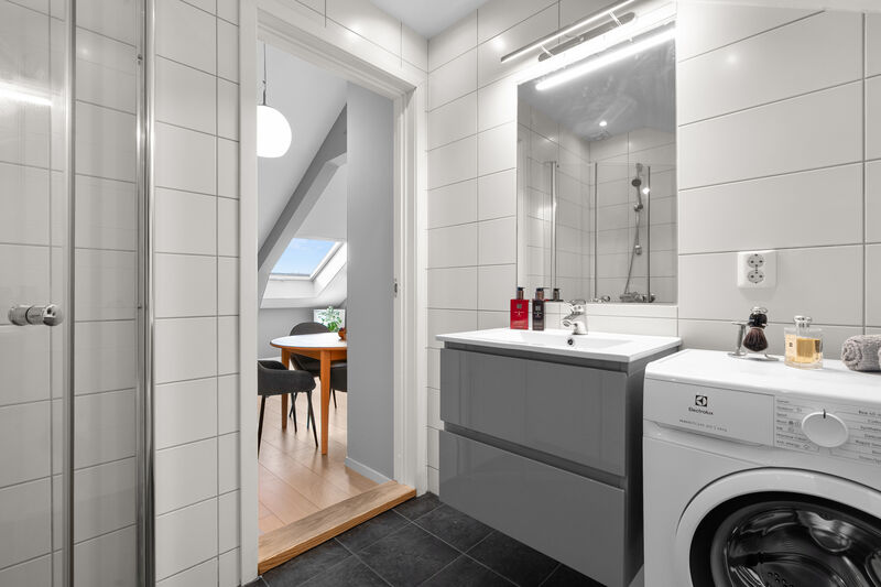 Moderne baderomsinnredning med skuffer og heldekkende servant, speil med belysning over. I hjørnet er det plass og opplegg for vaskemaskin.